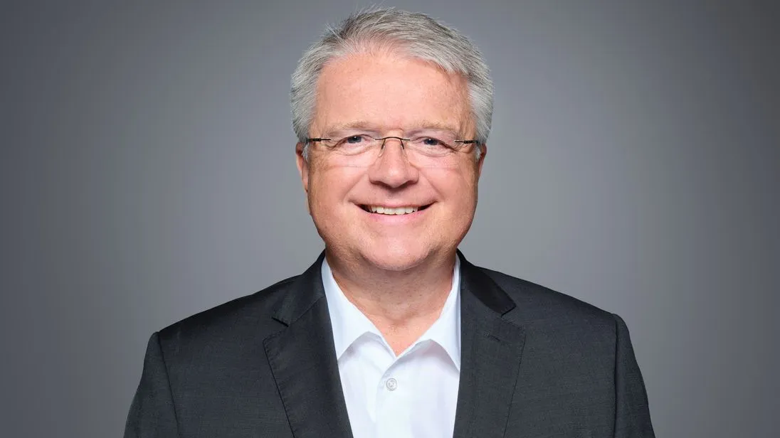 Ein weißer Mann in einem schwarzen Jackett und weißem Hemd vor grauem Hintergrund.